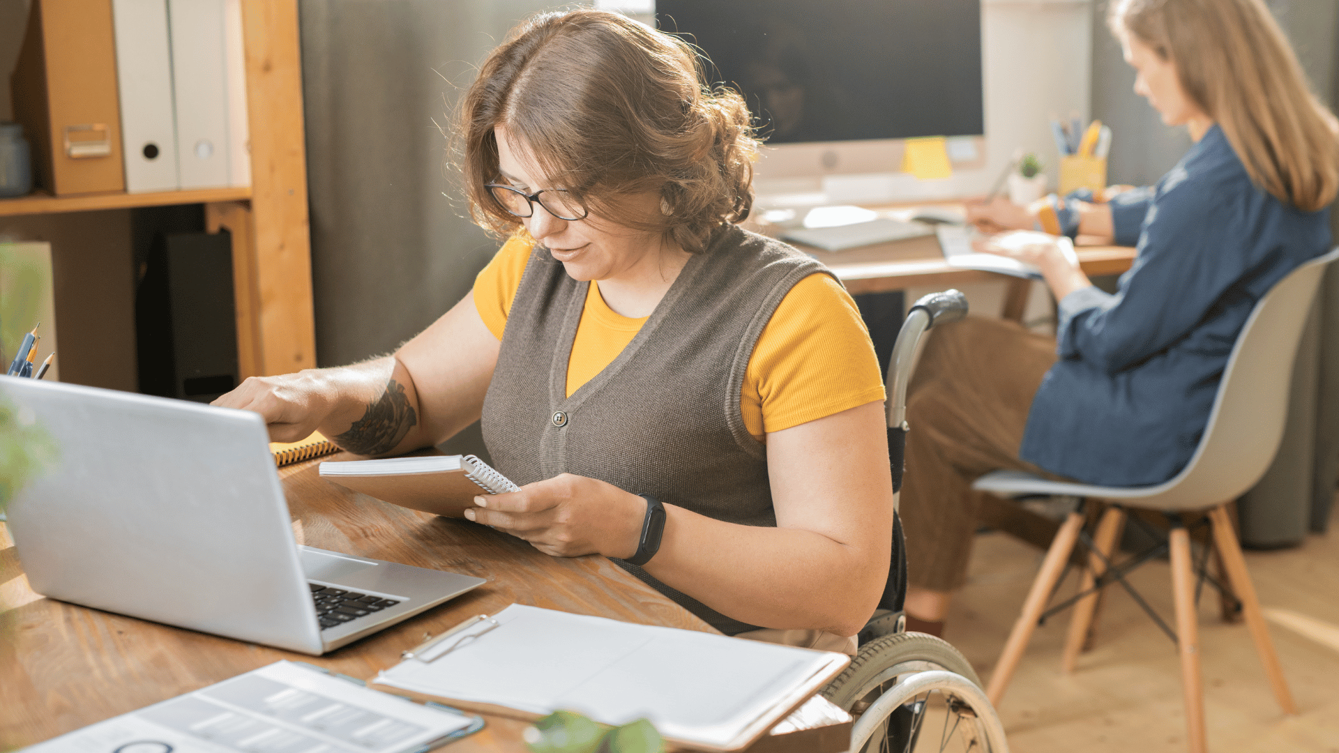 A woman in a wheelchair is sitting at her desk looking with a notebook in one hand and a computer open in front of her. Another woman is sitting at a desk in the background.