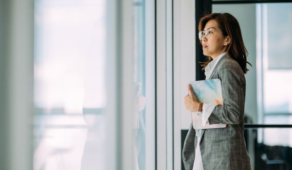 A woman wearing glasses and holding a tablet is standing and staring out the window in her office.