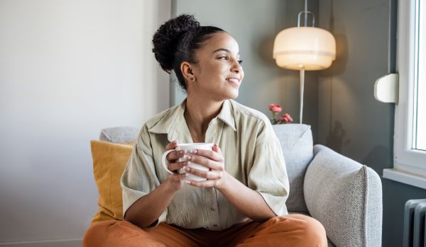 A young woman is sitting in a soft chair holding a cup of tea and smiling out the window
