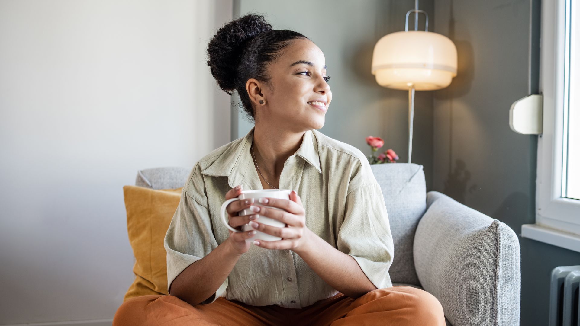 A young woman is sitting in a soft chair holding a cup of tea and smiling out the window