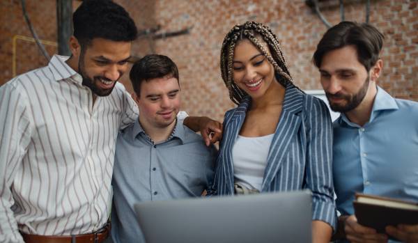 There are four employees in an office gathered around a laptop. They are smiling looking at the laptop.