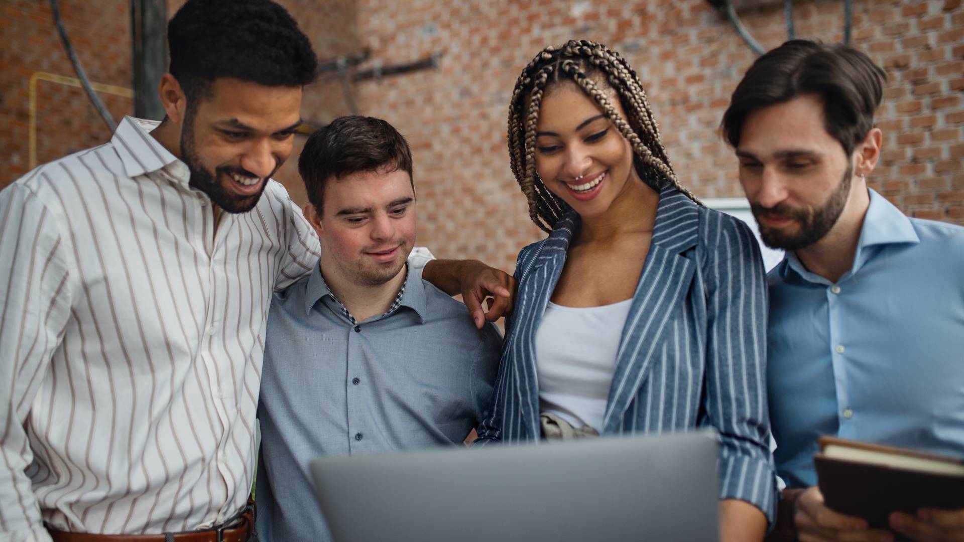 There are four employees in an office gathered around a laptop. They are smiling looking at the laptop.