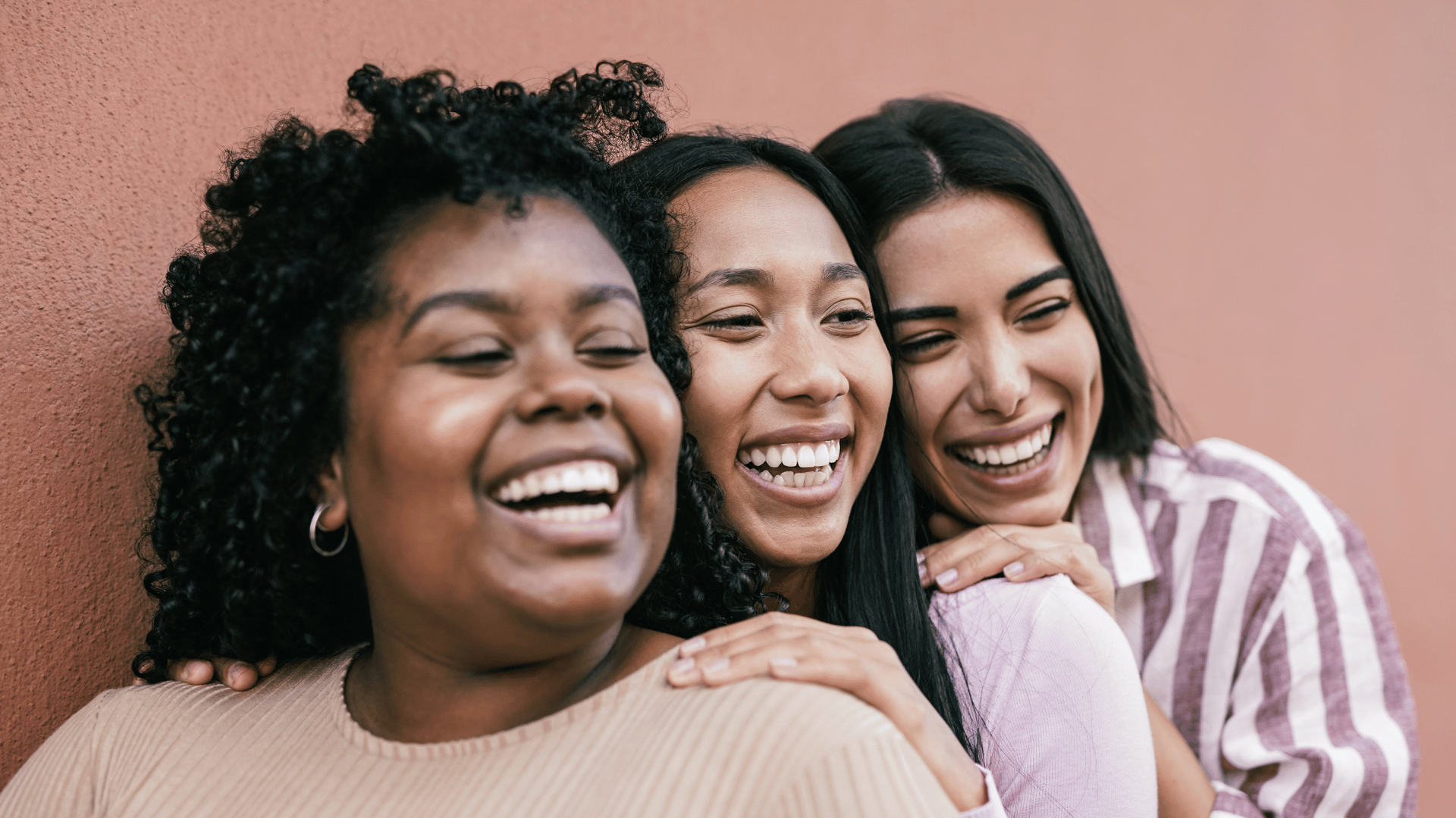 3 women of color standing near each other and smiling. We see them only from their shoulders up. A pinkish/tanish concrete wall is behind them.