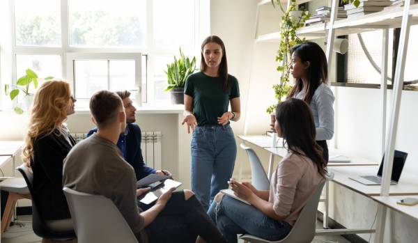 There is a team having a meeting. There are one people sitting on chairs in a circle and they are in front of two women who are standing. One of them is speaking to them.