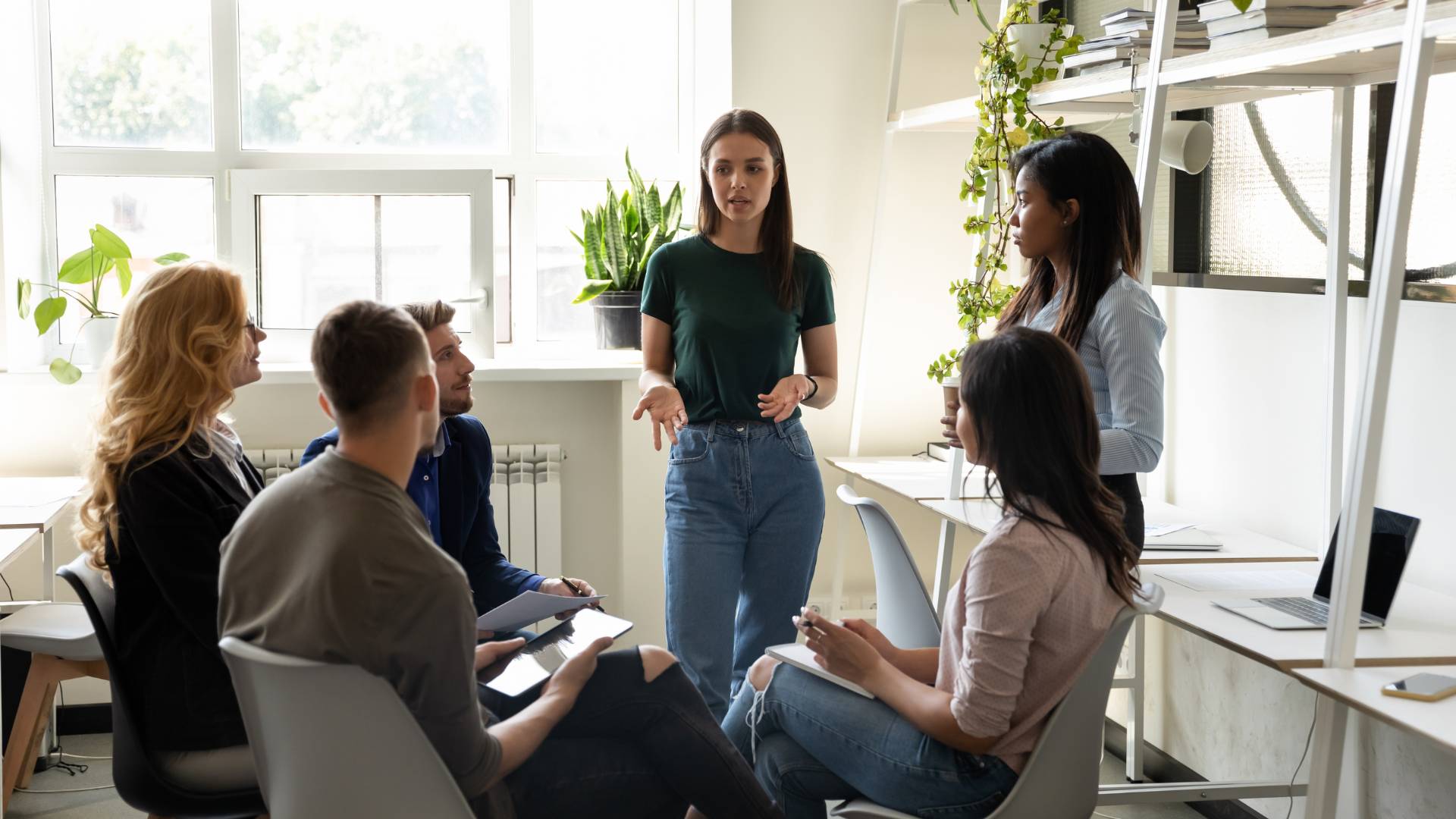There is a team having a meeting. There are one people sitting on chairs in a circle and they are in front of two women who are standing. One of them is speaking to them.