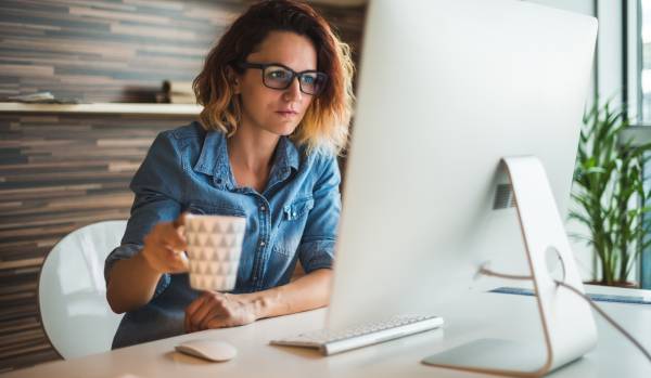 A woman with glasses is sitting at a desk holding a coffee mug. She is looking at her desktop.