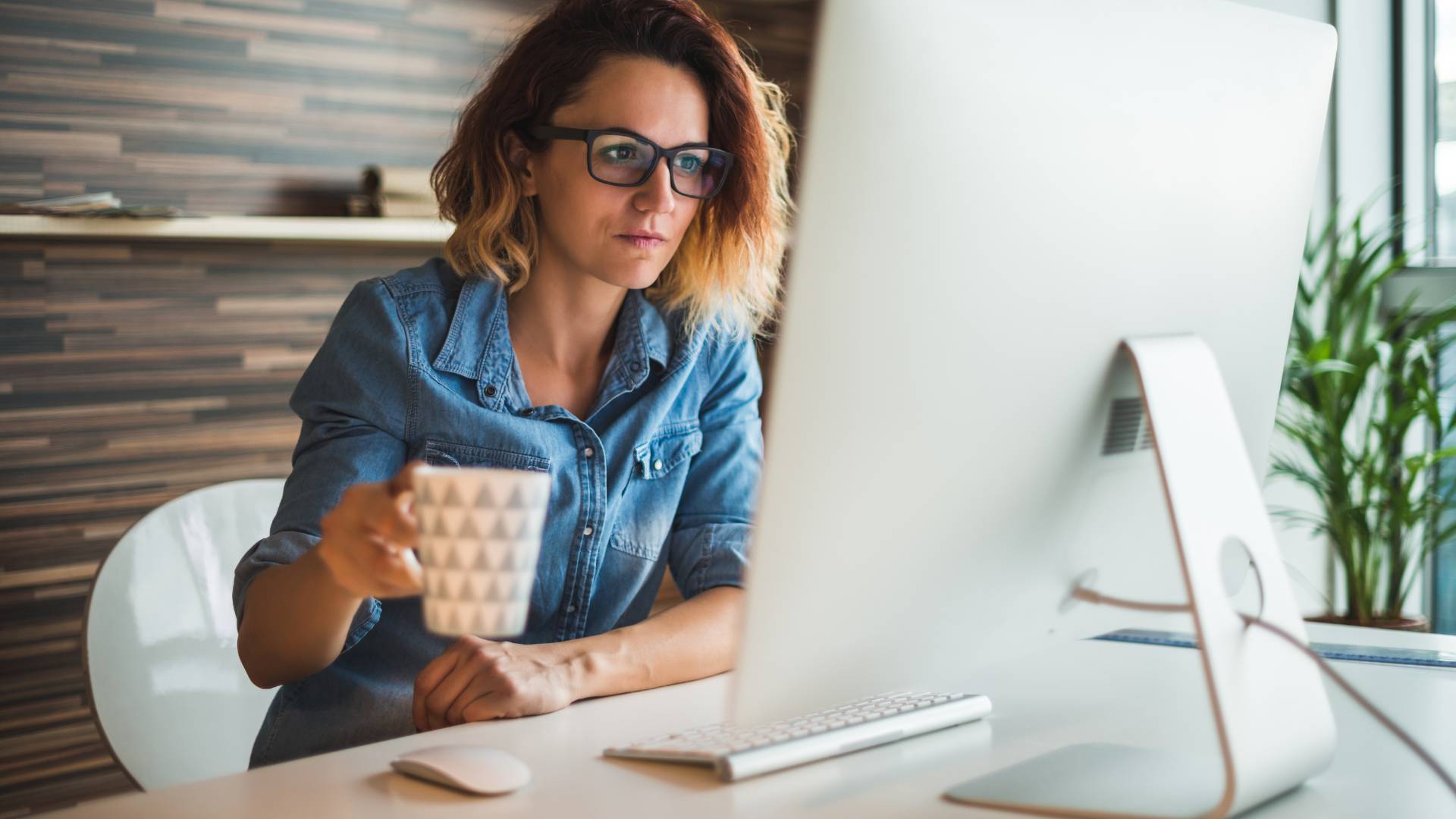 A woman with glasses is sitting at a desk holding a coffee mug. She is looking at her desktop.