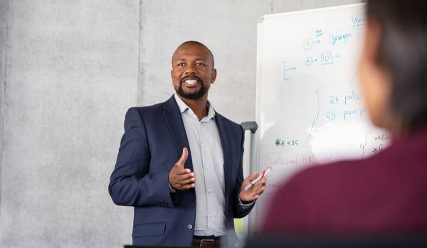 A man is standing at a whiteboard leading a training. We are looking over the shoulder of one of the participants.