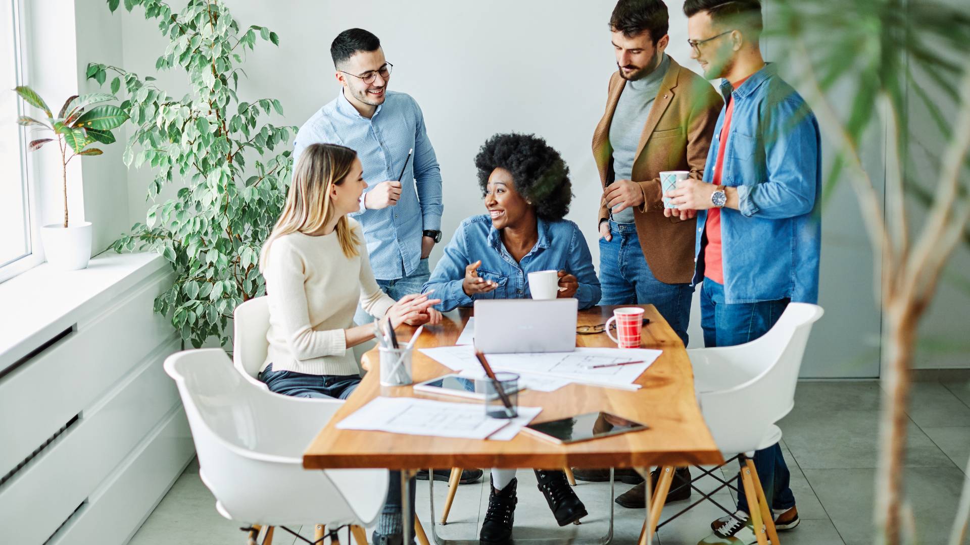 There's a team having a meeting at a table. There are three men standing, talking and laughing with two women who are sitting down also talking.