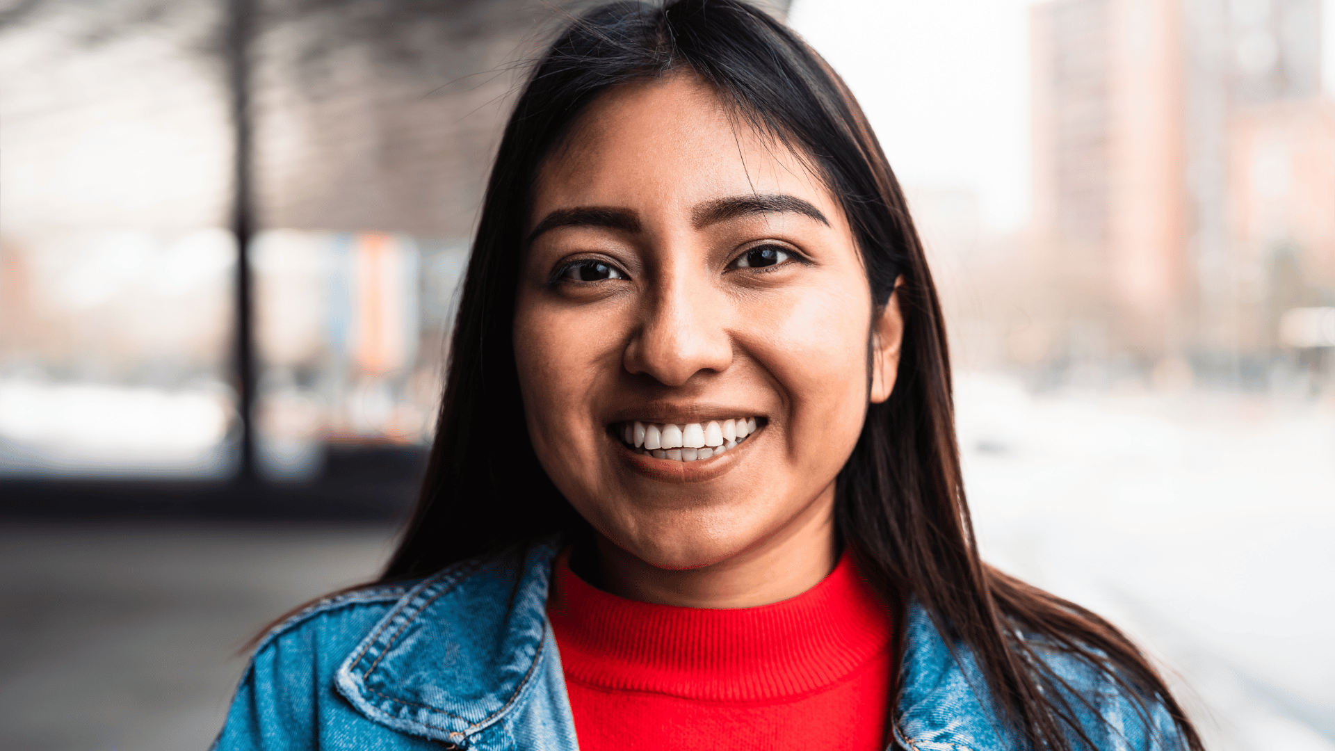 Native American woman smiling at the camera. She is wearing a red mock turtleneck and a denim jacket.
