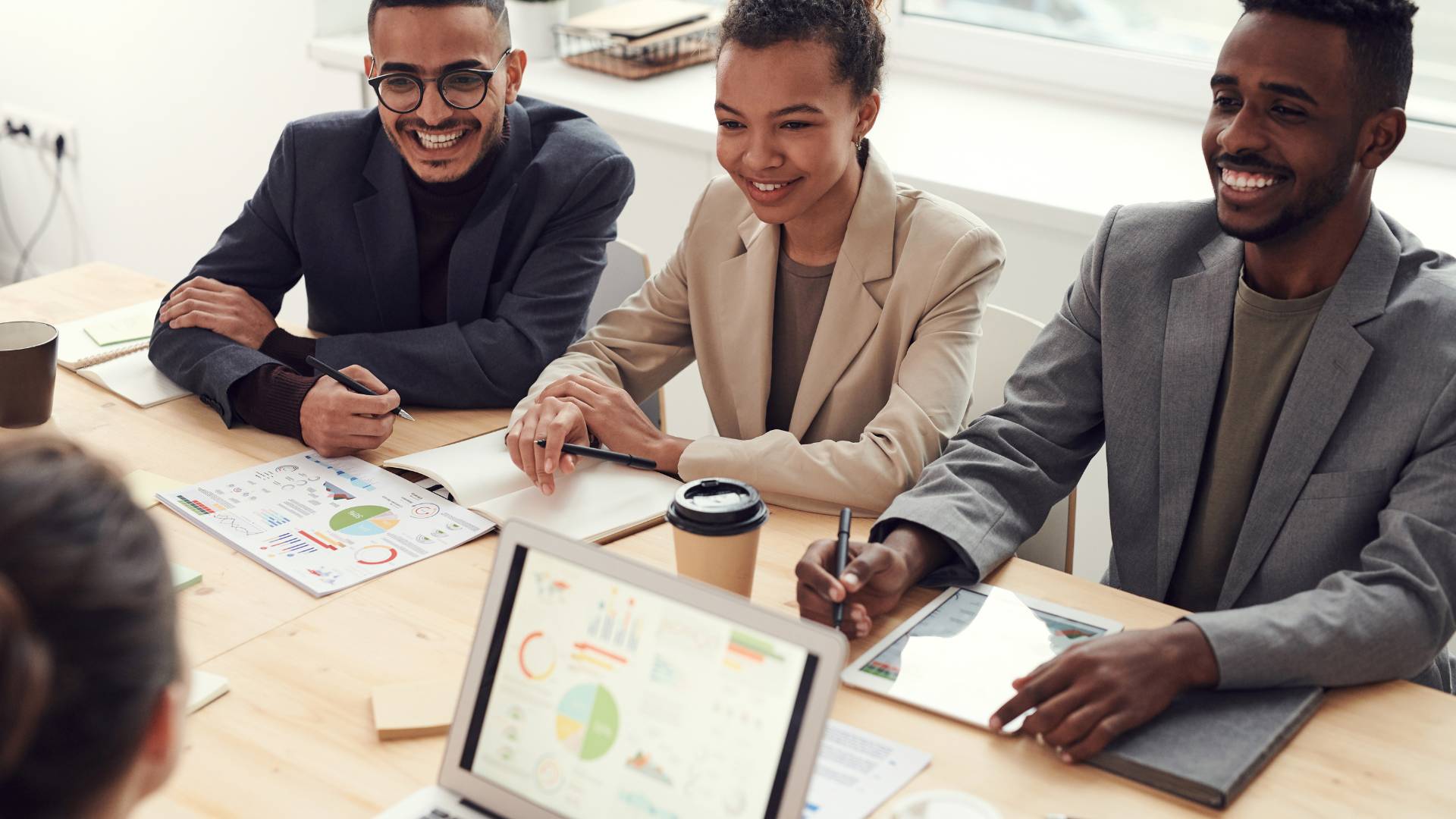 There are three employees sitting at a table together in the office engaging in a meeting with others. There's a laptop on the table with a chart displayed on the screen.