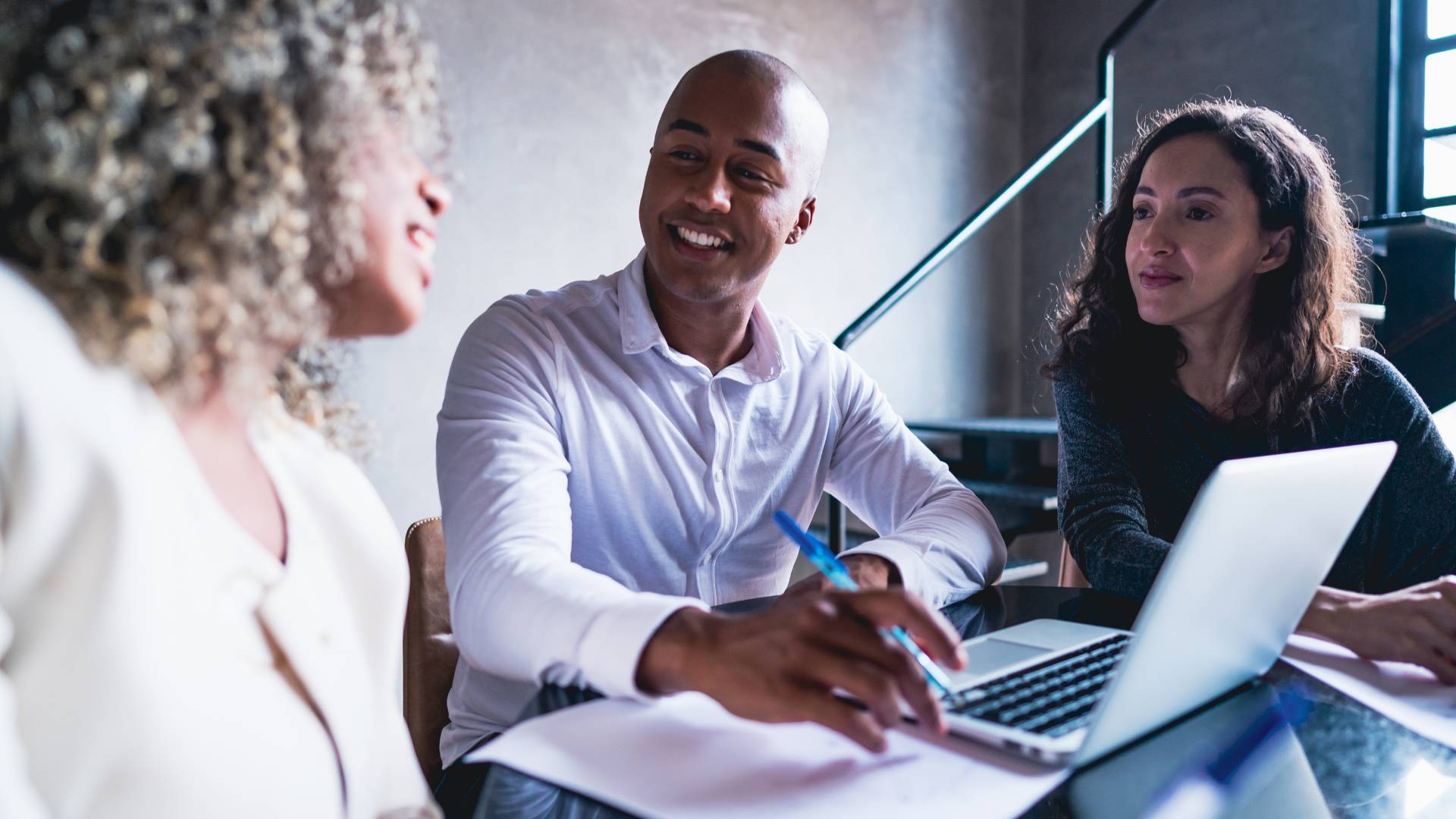 Three professionals are smiling and working together around a laptop.