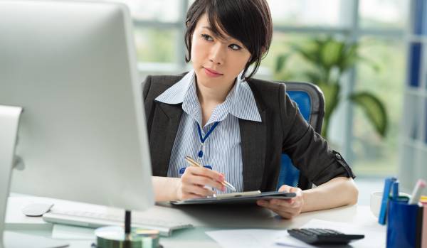 A woman is sitting at a desk in front of a computer. She's holding a stylus in her hand and a tablet as well.