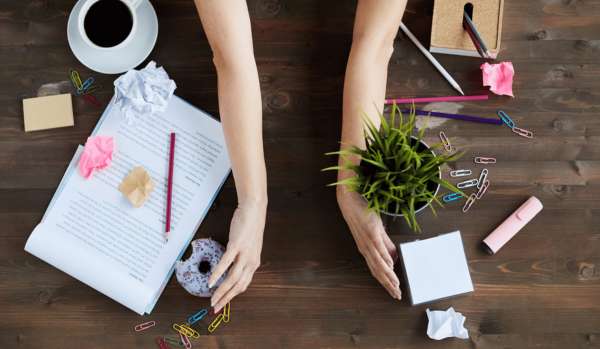 A messy desk being cleaned off. A woman’s arms are in the middle of the desk, facing outward, as if they’re about to sweep everything off to the sides of the desk.