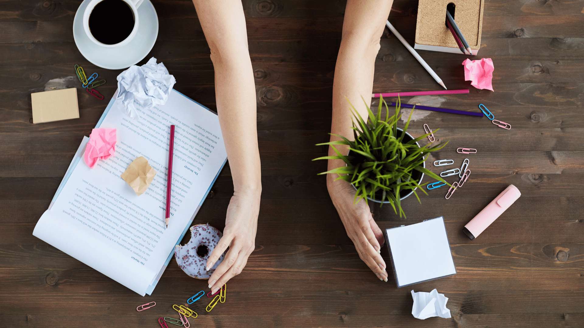 A messy desk being cleaned off. A woman’s arms are in the middle of the desk, facing outward, as if they’re about to sweep everything off to the sides of the desk.