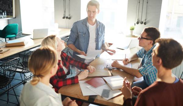 A group of workers are in an office gathered around a table brainstorming ideas. There are five people in total.