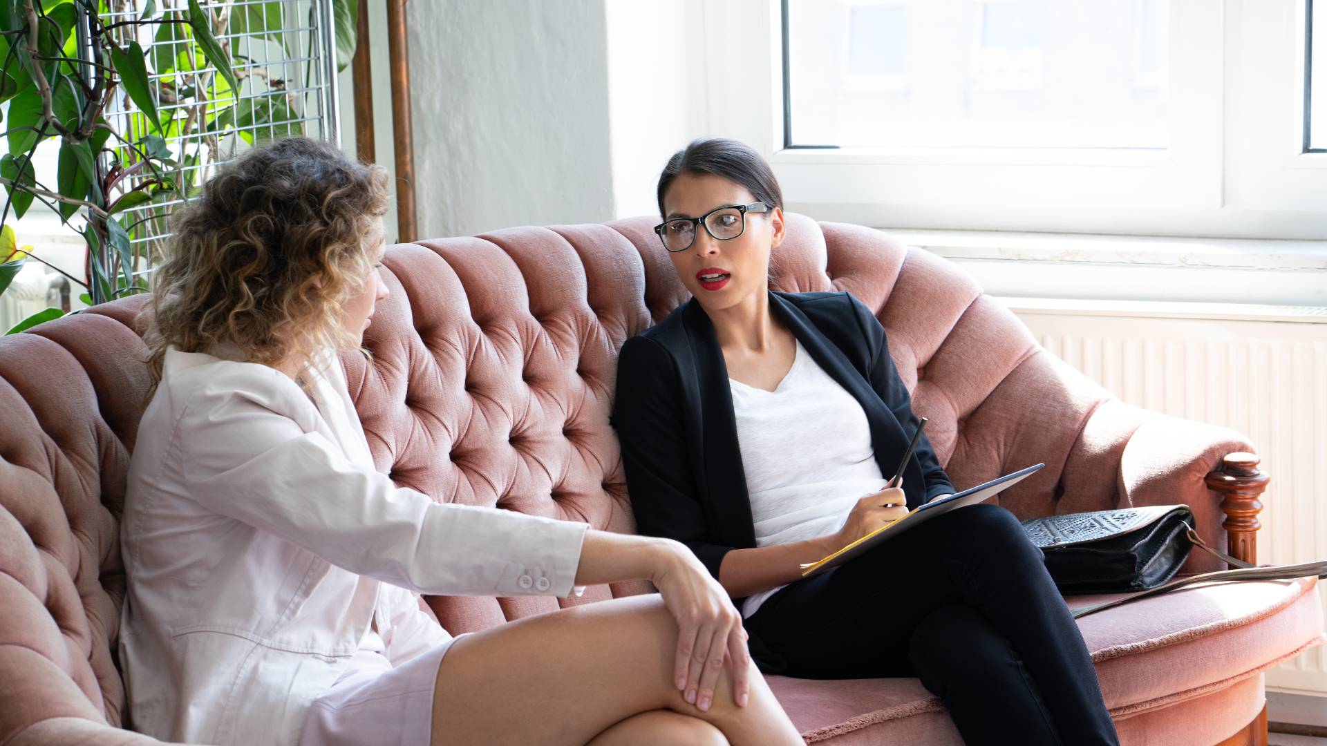 Two professional women are having a one-on-one meeting. They are sitting on a couch in an common area with plants and a window behind them.