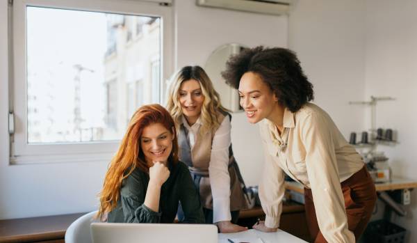 There are three women around a table smiling and looking at a laptop. They're in a meeting.