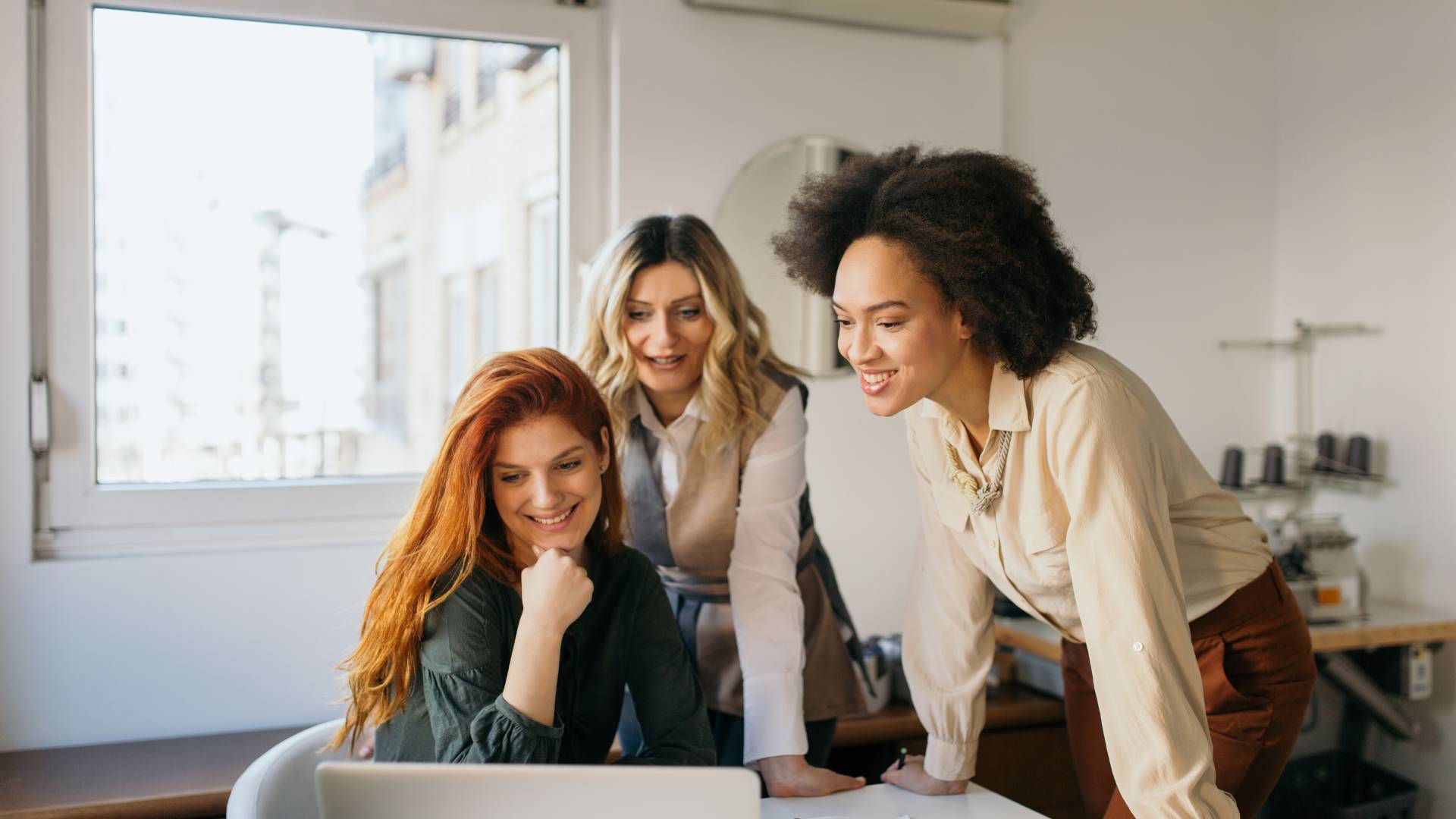 There are three women around a table smiling and looking at a laptop. They're in a meeting.