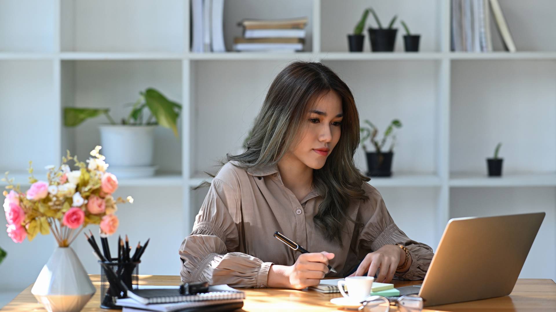 A young woman is looking at her laptop and taking notes with a pen and notebook.