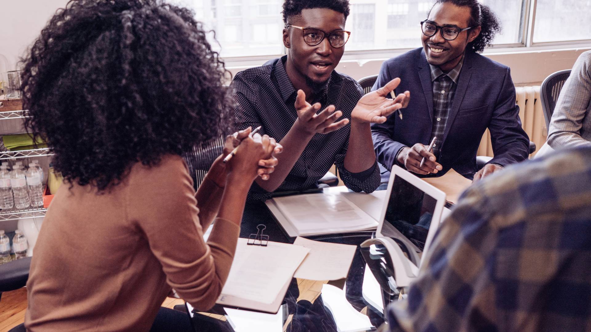 There are a group of workers sitting around a table in an office engaged in a meeting. Two of the workers are males and wearing glasses.