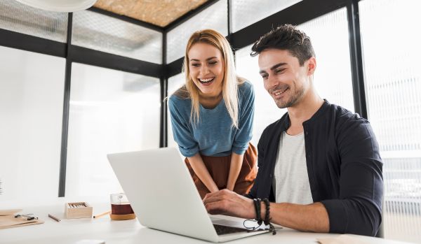 Two coworkers are smiling as they compare learning tools on one of their laptops.