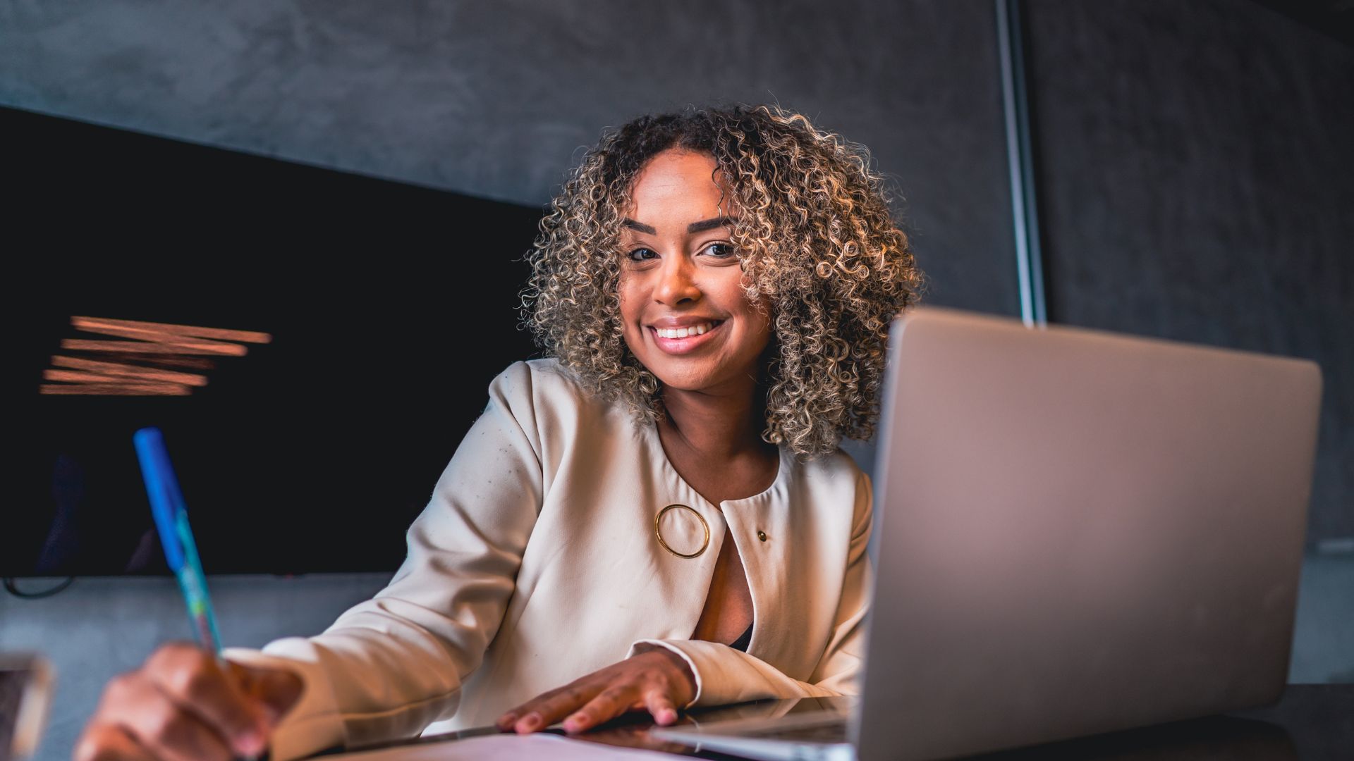 An HR leader is getting ready to launch a new manager training on her laptop. She is smiling at the camera while writing something down.