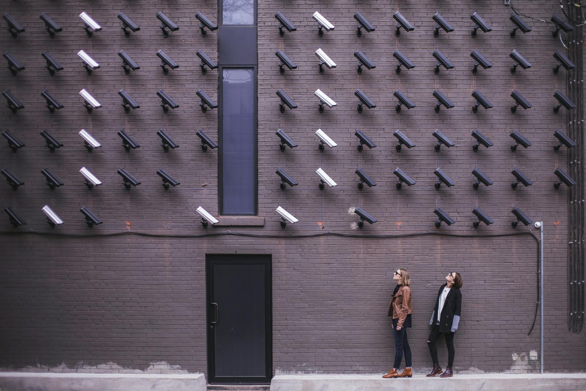 Two women standing against a brick wall covered with numerous security cameras, some white and some black, all pointing downward.