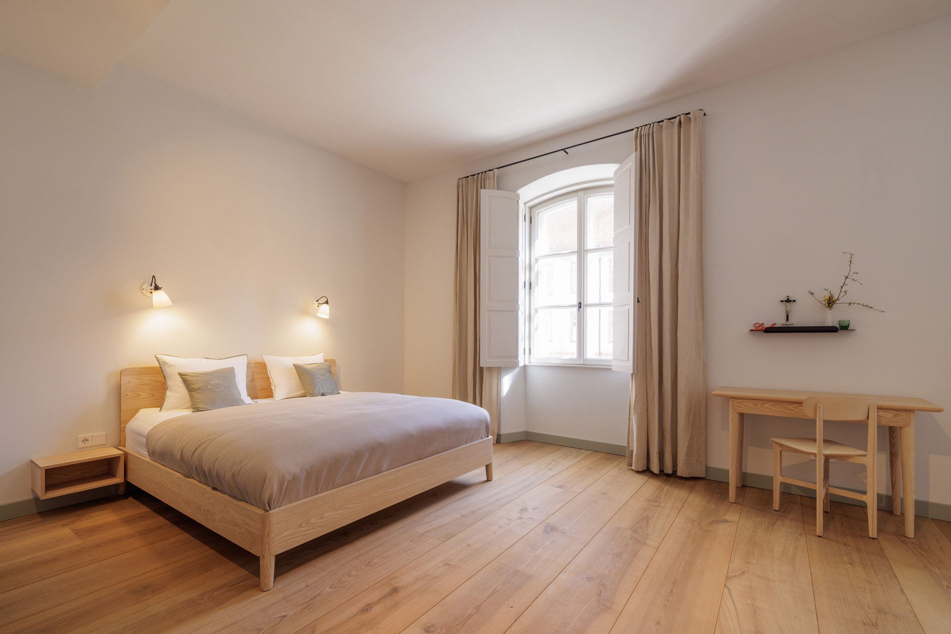 Frontansicht eines Gästezimmers mit Wandaltar und Tisch, Eichenparkett, Tischleuchte, Lounger und Fenstern mit langen Gardinen im Josephstrakt des Klosters Beuerberg, von formstelle gestaltet.