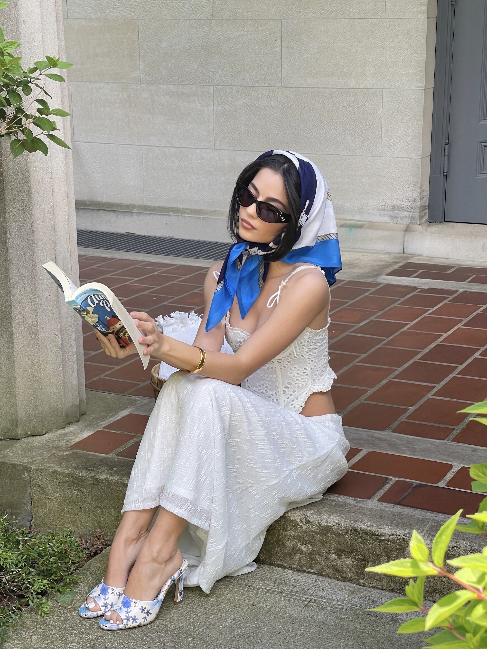 Woman in white eyelet dress with blue and white silk headscarf and sunglasses, sitting on stone ledge reading a book, wearing floral sandals in outdoor courtyard setting. Brought to you by Plural, NYC: Experts in Social Media Marketing Strategy and High-Quality Content Production.