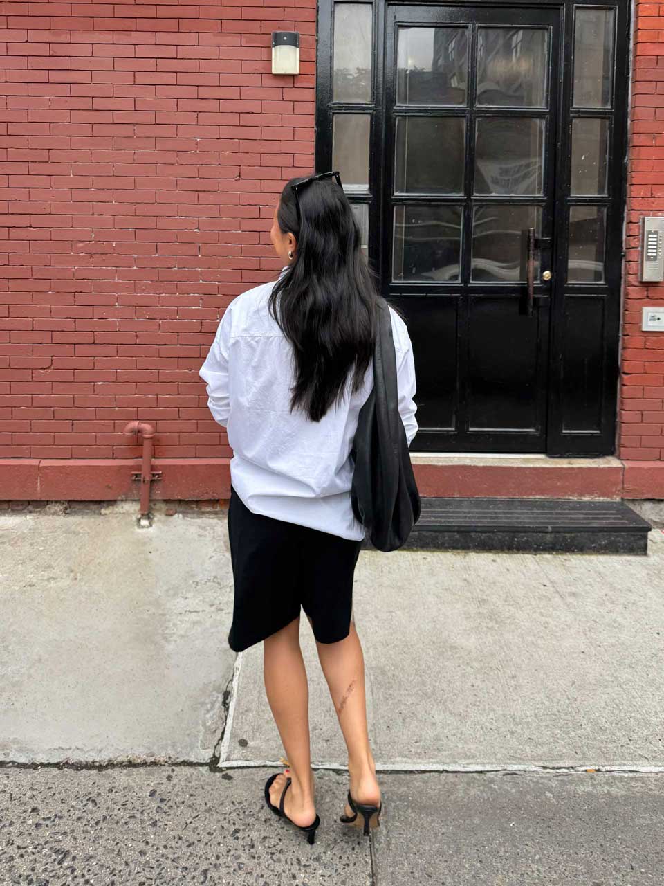 Woman with long black hair wearing a white shirt, black skirt, and black heeled sandals walking on a sidewalk by a brick building with a black door.