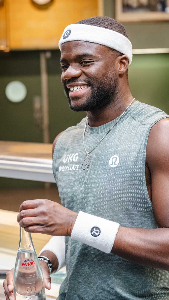 Smiling man in athletic wear holding a bottle of Evian water at an indoor setting.