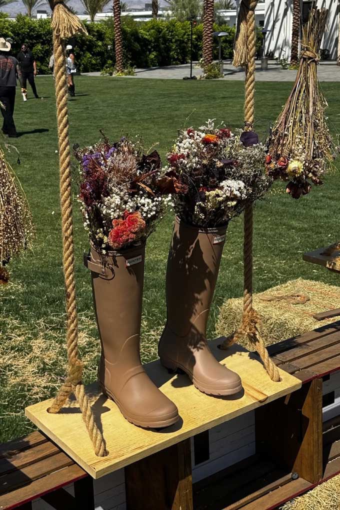 Tan rain boots filled with dried flowers suspended by rope on wooden swing with hay bales. Brought to you by Plural, NYC: Experts in Social Media Marketing Strategy and High-Quality Content Production.