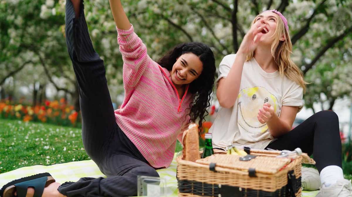 Two Aerie models laughing together on a picnic blanket, surrounded by blooming trees and tulips. One wears a pink knit sweater, the other a white t-shirt with a smiley face graphic, enjoying a sunny day outdoors with a picnic basket.