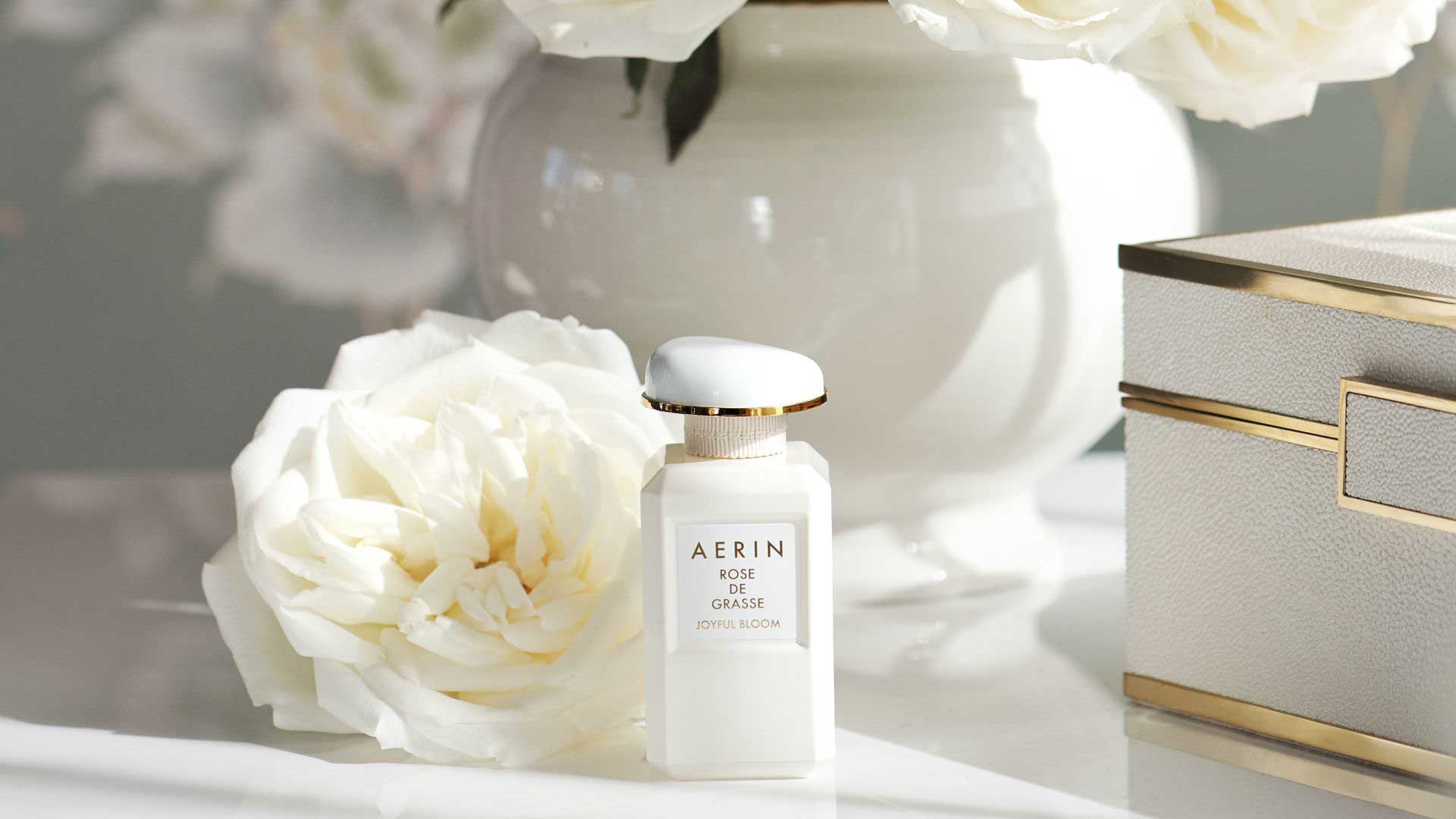 AERIN Rose de Grasse Joyful Bloom perfume bottle next to a white rose and a textured white box with gold trim.