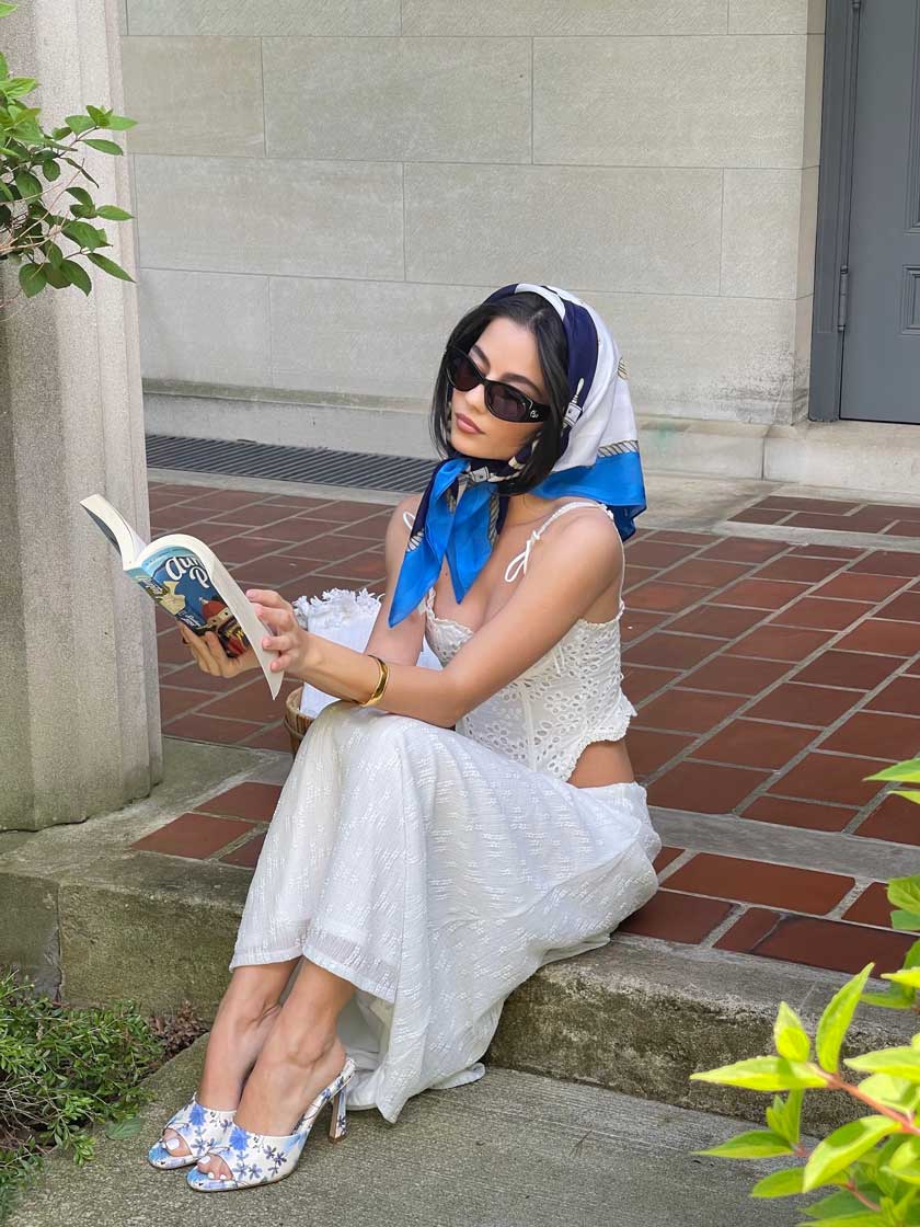 Woman in white eyelet dress with blue and white silk headscarf and sunglasses, sitting on stone ledge reading a book, wearing floral sandals in outdoor courtyard setting. Brought to you by Plural, NYC: Experts in Social Media Marketing Strategy and High-Quality Content Production.