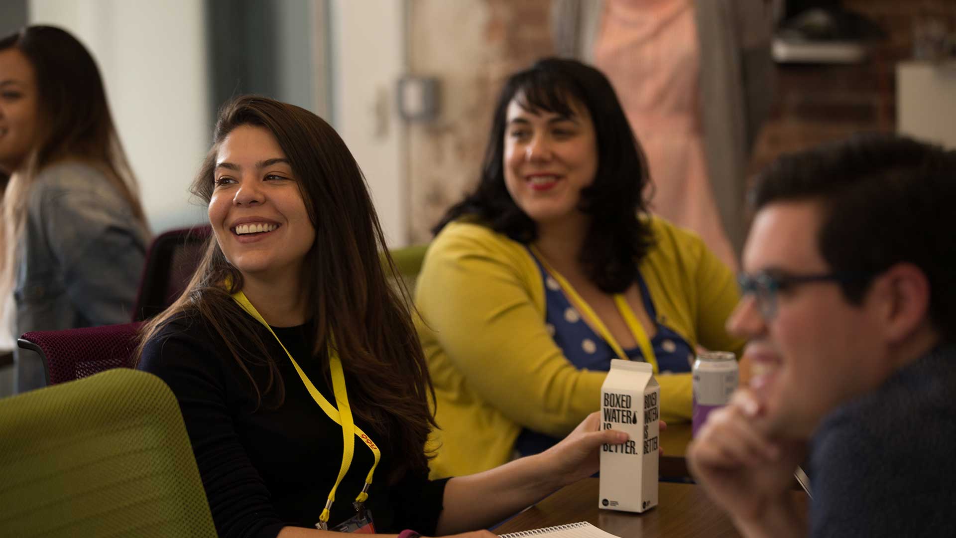 Smiling woman with a yellow lanyard holding a carton labeled 'Boxed Water Is Better' sitting at a table with other people in a casual indoor setting.
