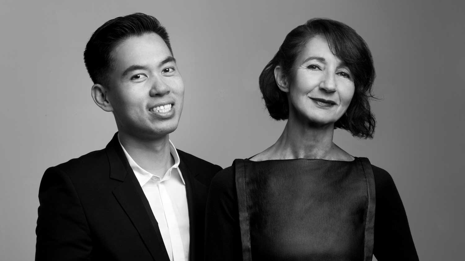 Black and white portrait of a smiling young man in a suit and an older woman in a dark dress, standing side by side.