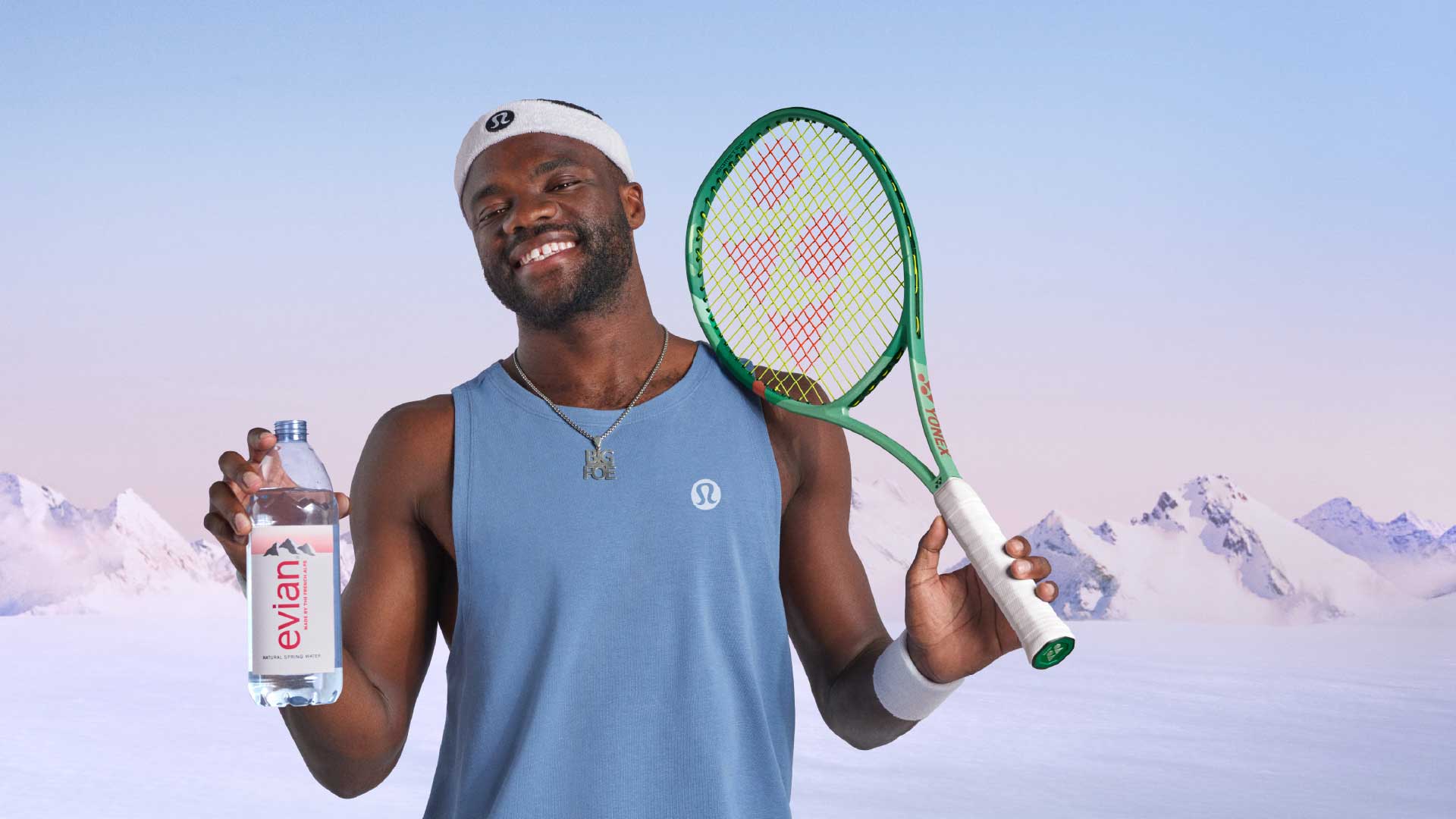 Frances Tiafoe in blue Lululemon tank top and white headband holds a green tennis racket and a bottle of Evian water against snowy mountain background.