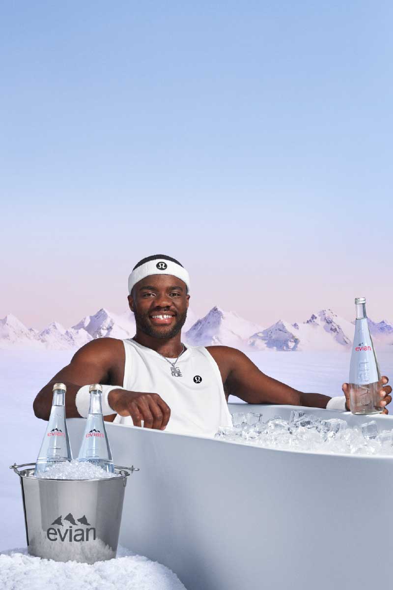 Frances Tiafoe in white athletic wear and headband sitting in an ice-filled tub holding a bottle of Evian water with snow mountains in the background.