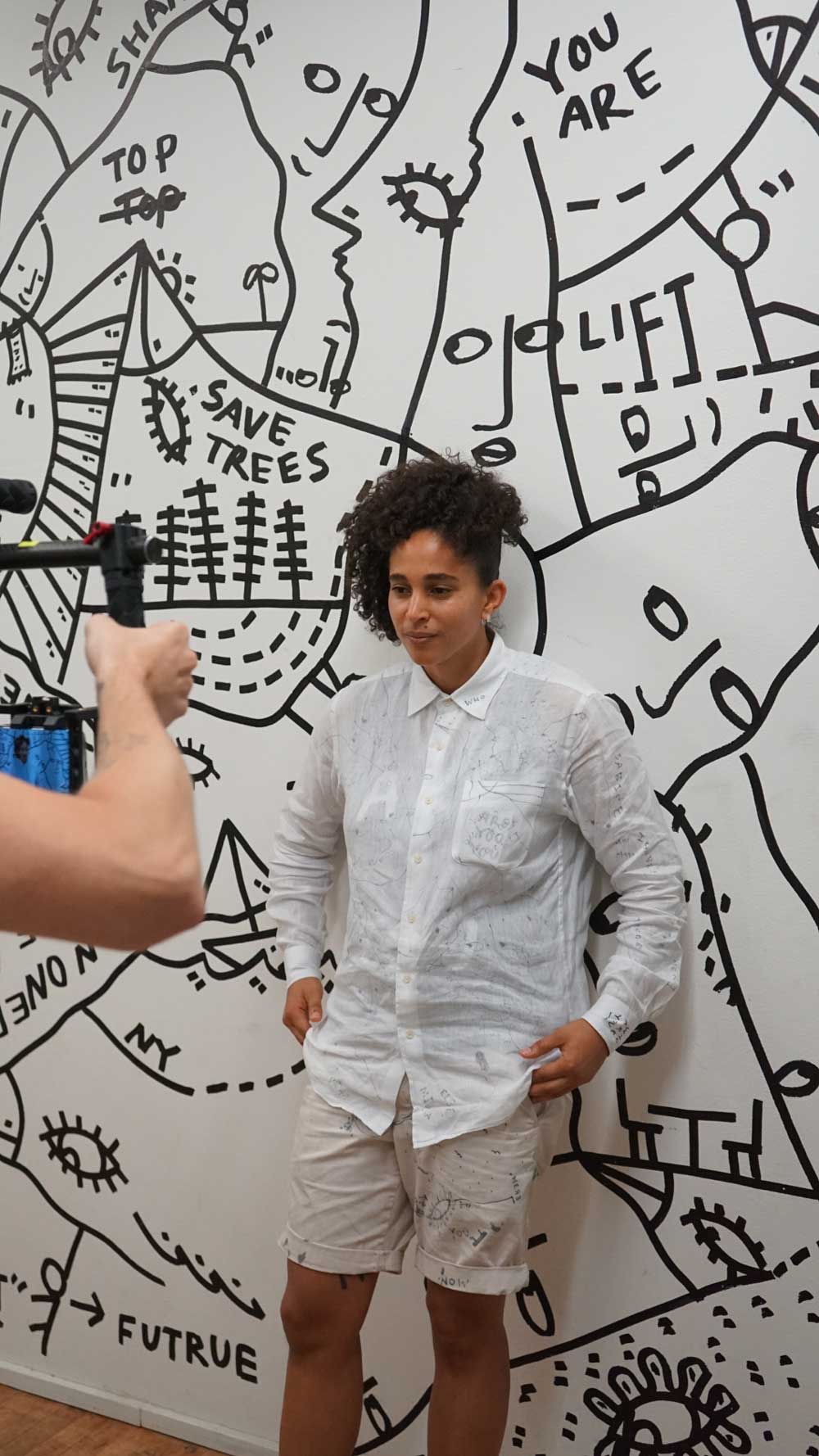 Shantell Martin stands in front of a black-and-white mural while being filmed by a camera operator. Brought to you by Plural, NYC: Experts in Social Media Marketing Strategy and High-Quality Content Production.