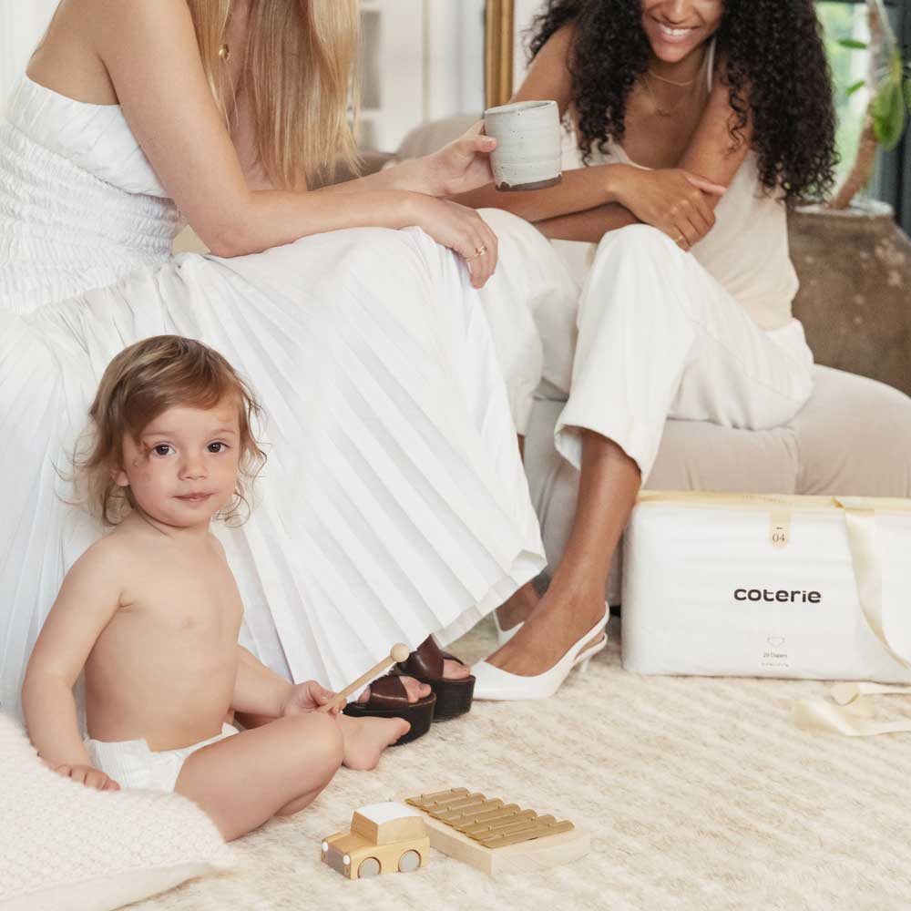 Two women in white outfits looking at a baby in a diaper. Brought to you by Plural, NYC: Experts in Social Media Marketing Strategy and High-Quality Content Production.