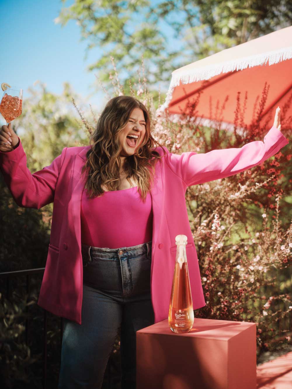 Woman in bright pink jacket and top holding a cocktail glass outdoors with a bottle on a pedestal nearby.