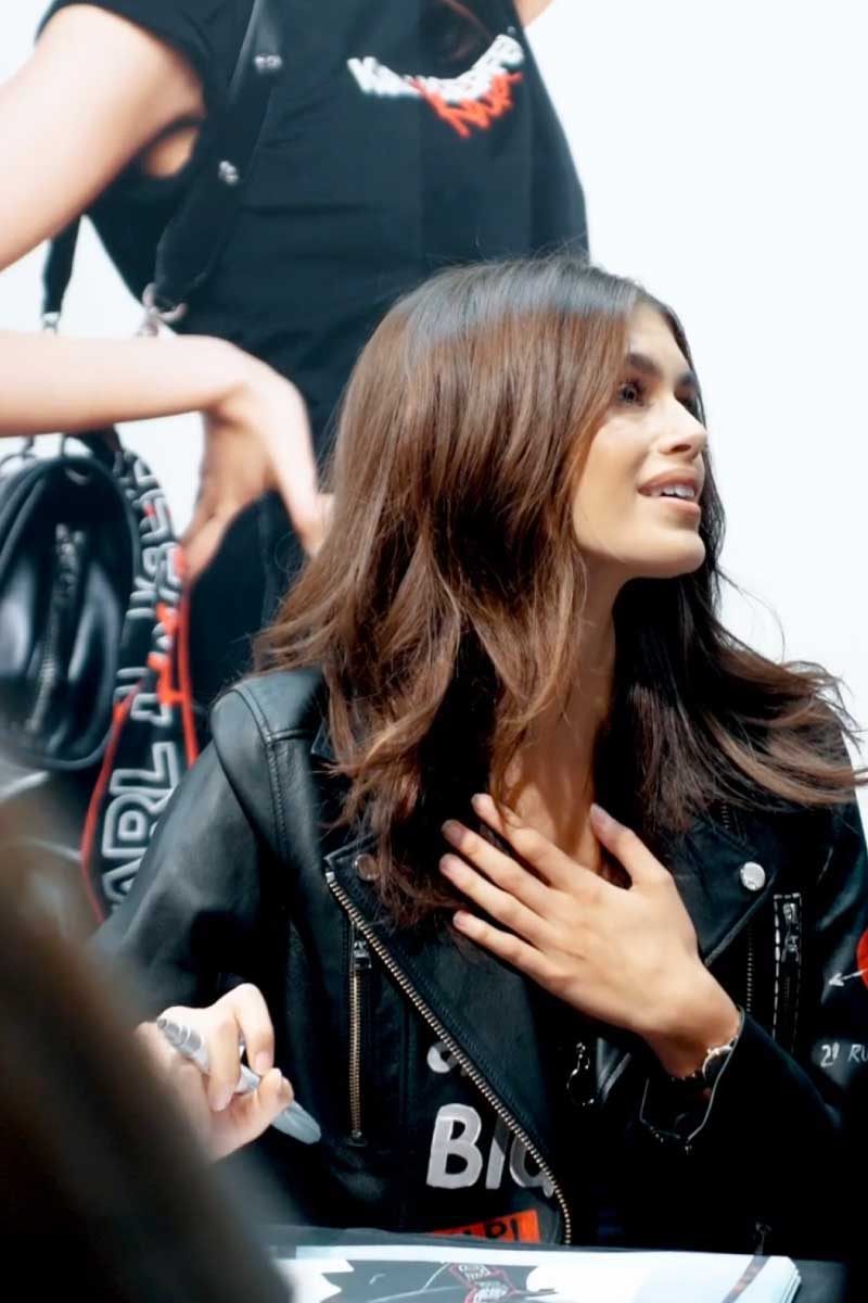 Kaia Jordan Gerber wearing a black leather jacket, holding a pen and placing her hand on her chest while looking to the right.