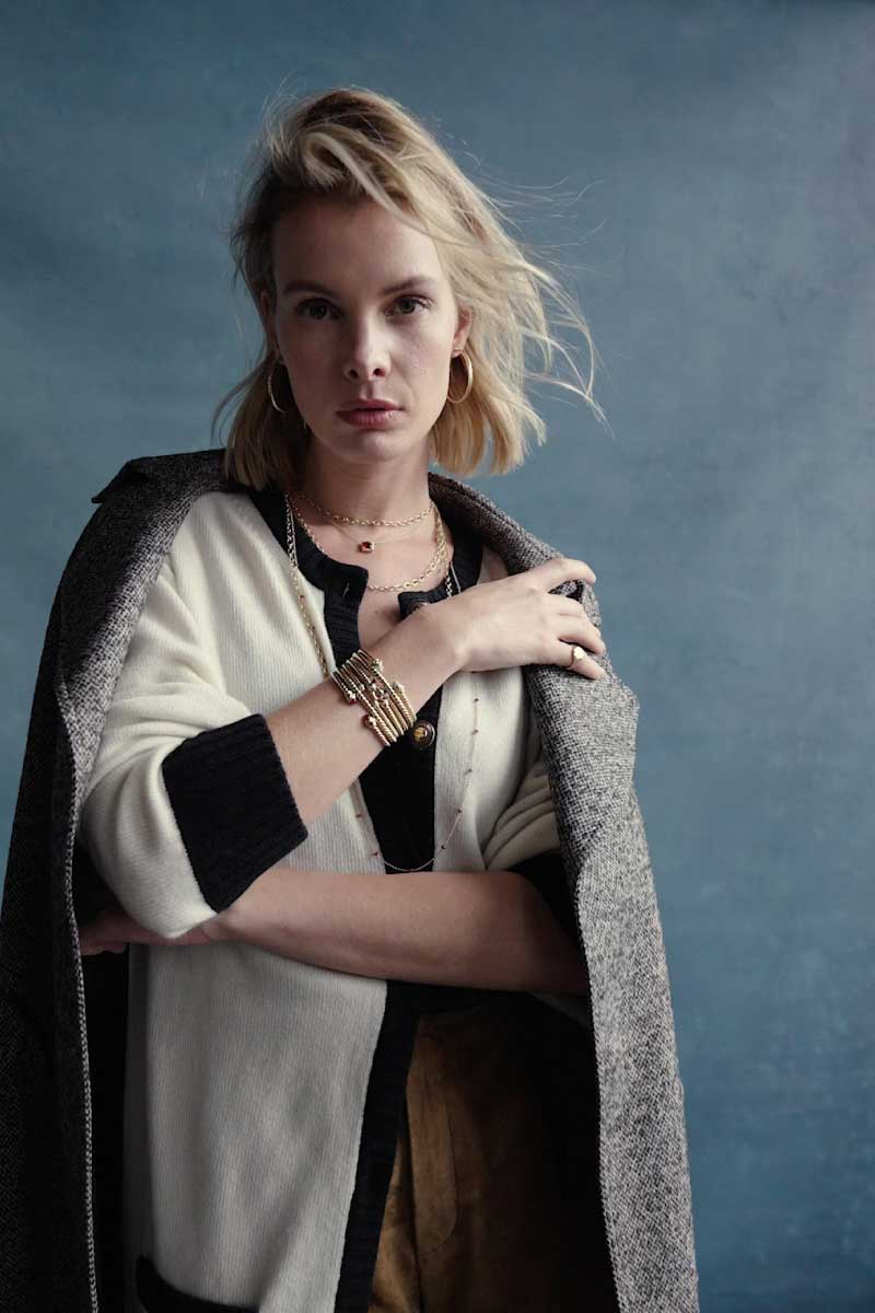 Blonde woman wearing a black and white cardigan, David Yurman gold bracelets, and hoop earrings, with a gray coat draped over her shoulders against a blue background.