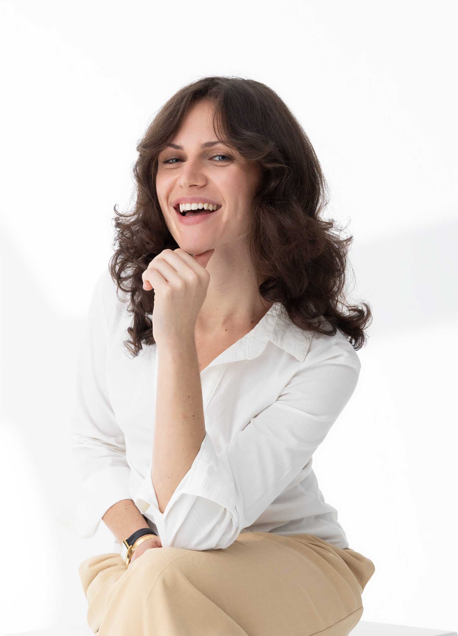 Smiling woman with curly dark hair wearing a white shirt and beige pants, resting her chin on her hand against a white background.