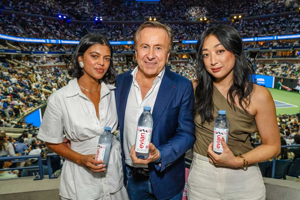 Three people standing at a tennis stadium holding Evian water bottles, with a tennis match and crowd in the background.