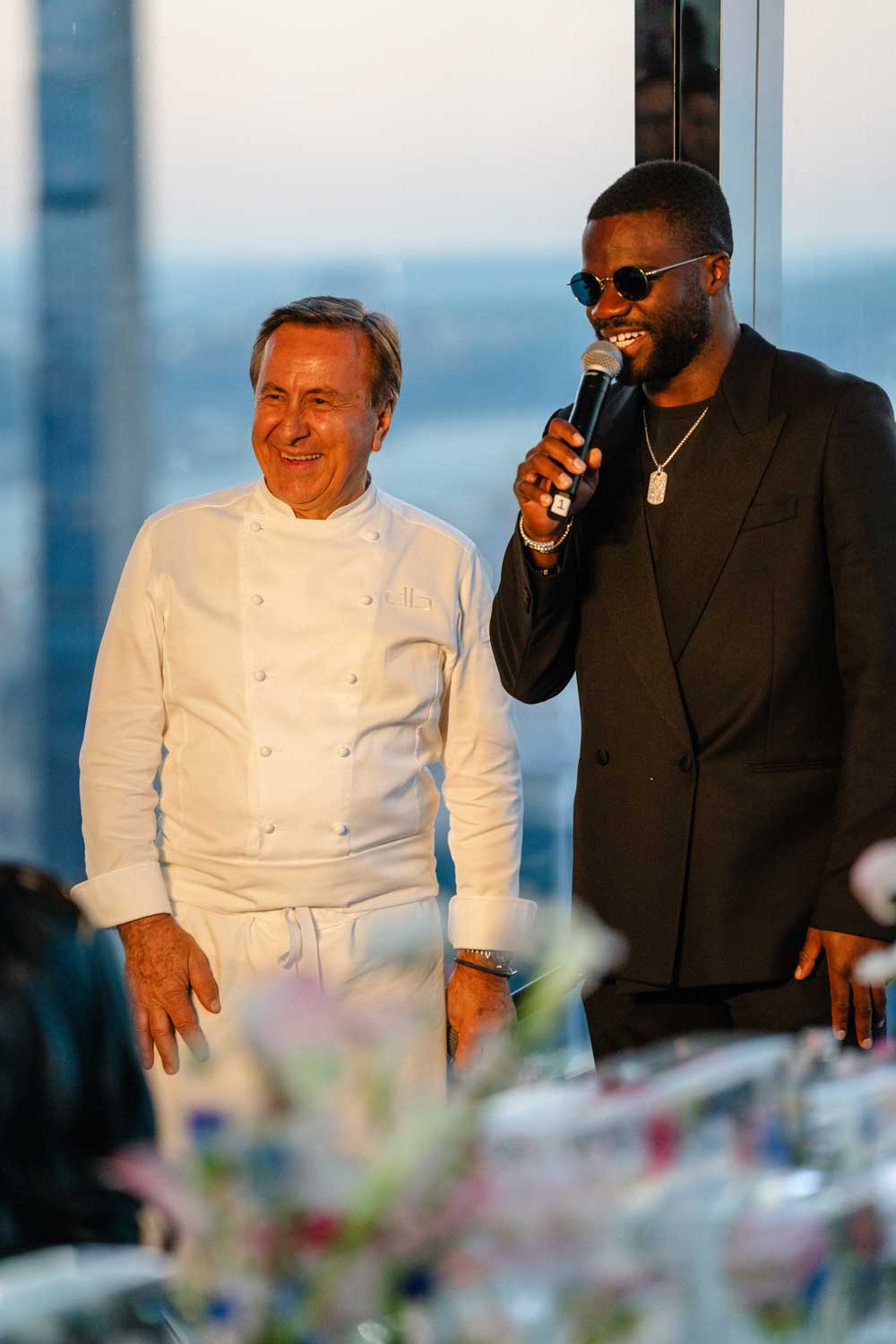 Man in white chef coat smiling next to man wearing sunglasses and black suit holding a microphone.