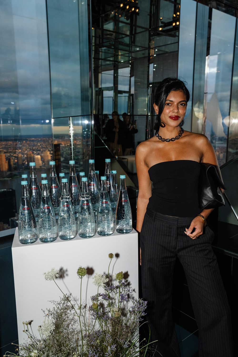 Woman in black strapless top and pinstripe pants standing next to a display of Evian water bottles and flowers.