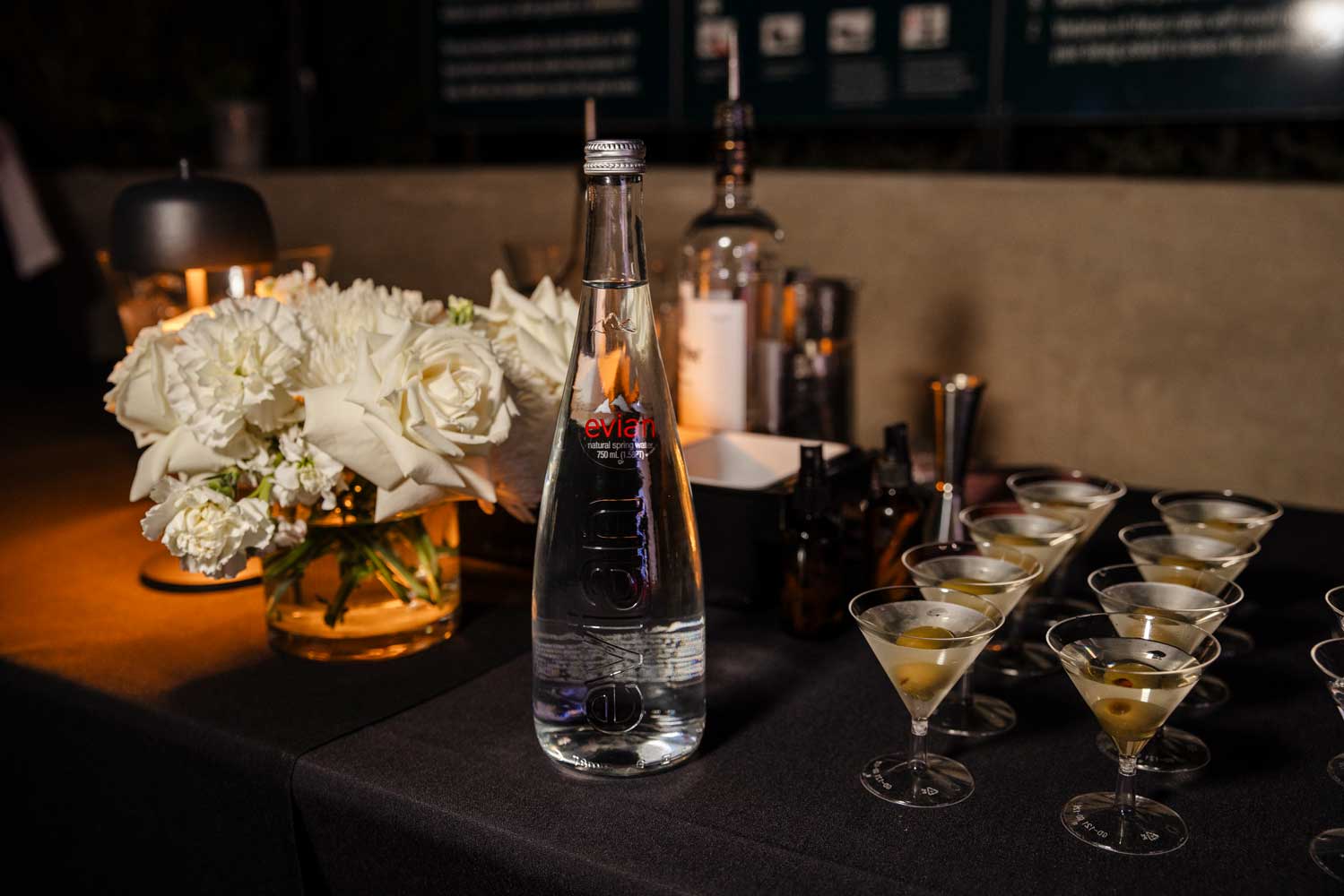 Glass Evian water bottle on table with white flowers and martini glasses with olives.